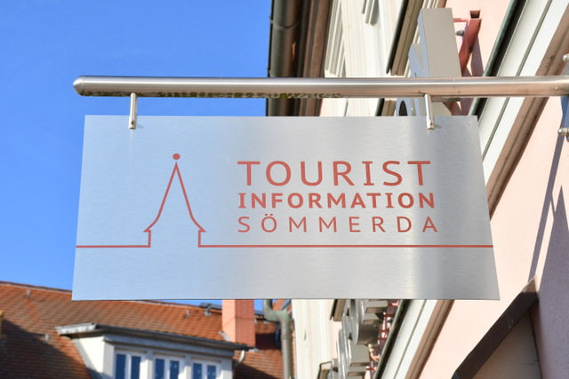 Ein Edelstahlschild hängt als Ausleger am Gebäude der Tourist-Information Sömmerda. Auf dem Schild stehen in Rot eine stilisierte Stadtsilhouette und der Schriftzug Tourist-Information Sömmerda. und der S