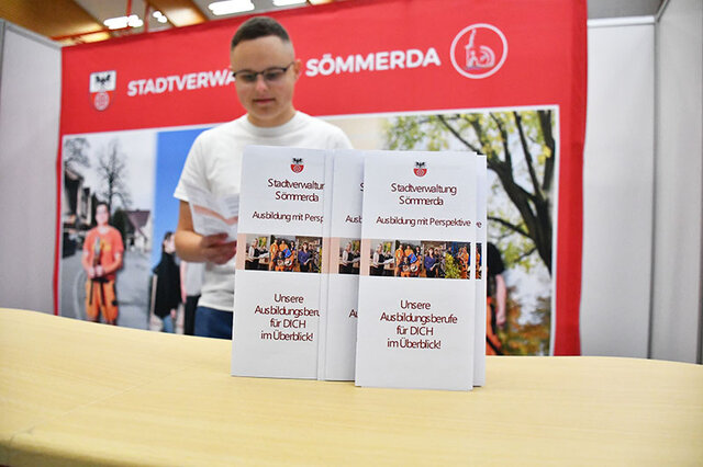 Vor einem großen Plakat der Stadt Sömmerda steht ein Counter aus hellem Holz. Auf diesem sind zwei Flyer hochkant aufgestellt. In ihnen werden Ausbildungsberufe bei der Stadt Sömmerda vorgestellt. Hinter dem Counter steht ein junger Mann, im weißen T-Shirt und mit Brille ud blättert durch einen der Flyer.