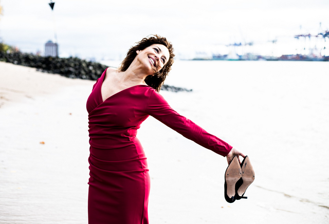 Isabel Varell Isabel Varell lächelnd in einem roten Kleid mit ihren Schuhen in der Hand an einem Strand