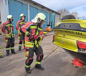 Ein Feuerwehrmann arbeitet mit Technik an einem auf dem Dach liegenden Pkw, während zwei weitere Feuerwehrleute zuschauen.