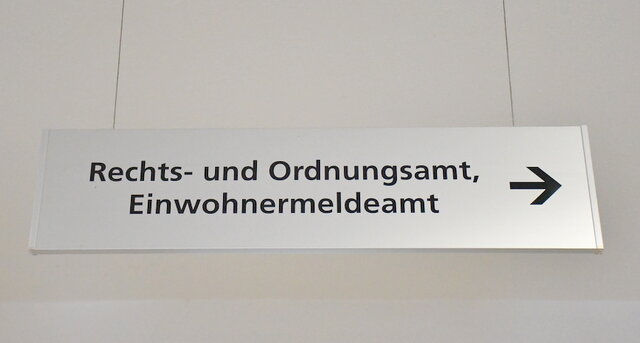 Von der Decke hängendes, rechteckiges Hinweisschild aus gebürstetem Edelstahl im Verwaltungsgebäude Poststraße. In der oberen Zeile steht Rechts- und Ordnungsamt, darunter das Wort "Einwohnermeldeamt".