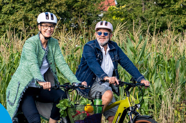 Zwei Personen mit Fahrradhelmen sitzen lächelnd auf Fahrrädern vor einem grünen Feld und werben für klimafreundliche Mobilität beim STADTRADELN in Sömmerda.