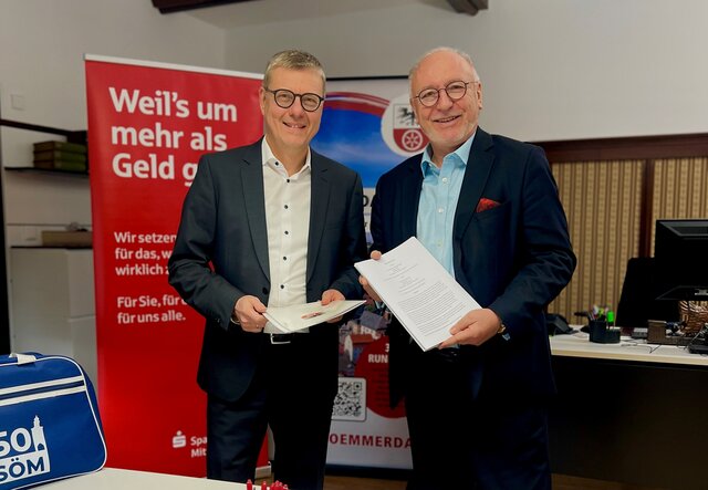 Bürgermeister Ralf Hauboldt und Sparkassen-Vorstandsvorsitzender Hans-Georg Dorst stehen im Rathaus nebeneinander und halten den frisch unterzeichneten Sponsoringvertrag für das Stadtjubiläum 2026 in den Händen. Im Hintergrund sind Werbebanner der Sparkasse und der Stadt zu sehen.