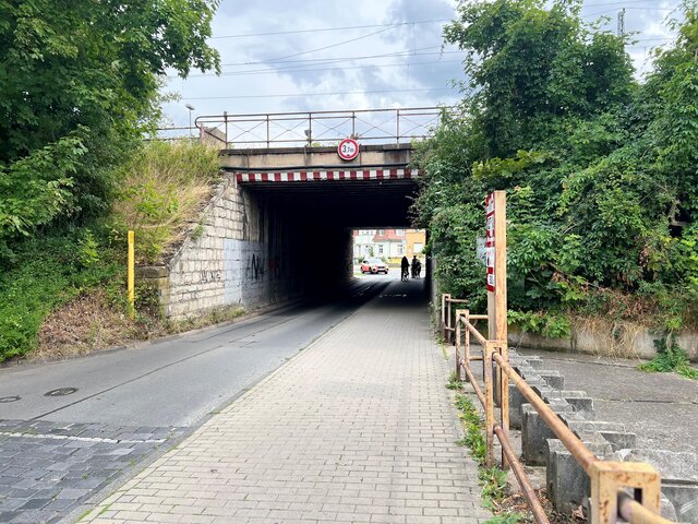 Blick auf den Bahnhofstunnel in Sömmerda mit Fahrbahn und Gehweg. Der Tunnel ist von dichter Vegetation umgeben, im Hintergrund sind Personen und Fahrzeuge zu sehen. Ein Schild weist auf die maximale Durchfahrtshöhe hin.