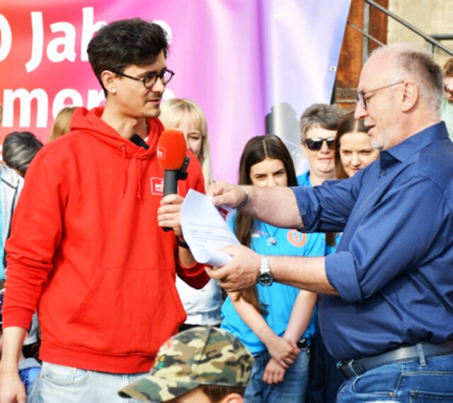 Ein Reporter von Antenne Thüringen hat Bürgermeister Ralf Hauboldt die Papierrolle mit der Aufgabe für den Wettbewerb übergeben. Sömmerdas Bürgermeister hält die Rolle in den Händen und liest die Aufgabe. Im Hintergrund hören Kinder erwartungsvoll zu.