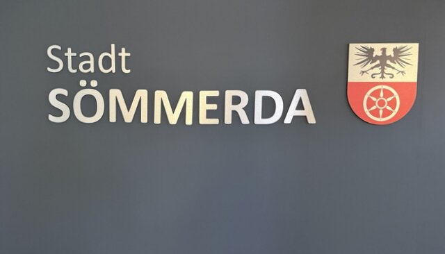 Element des Empfangstresens im Foyer des Sömmerdaer Rathauses. Ein grauer Hintergrund. Auf diesem steht in weißer Schrift zweilig Stadt Sömmerda. Daneben ist das Stadtwappen von Sömmerda angebracht.