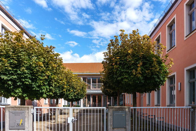 Die Musikschule in SÖmmerda von außen. Es ist ein u-förmiger Gebäudekomplex in Dunkelrosa. Vom Eingang von der Straßenseite her blickt direkt auf den Haupteingang der Musikschule. Im Innenhof der Musikschule stehen links und rechts des Hauptweges Bäume.