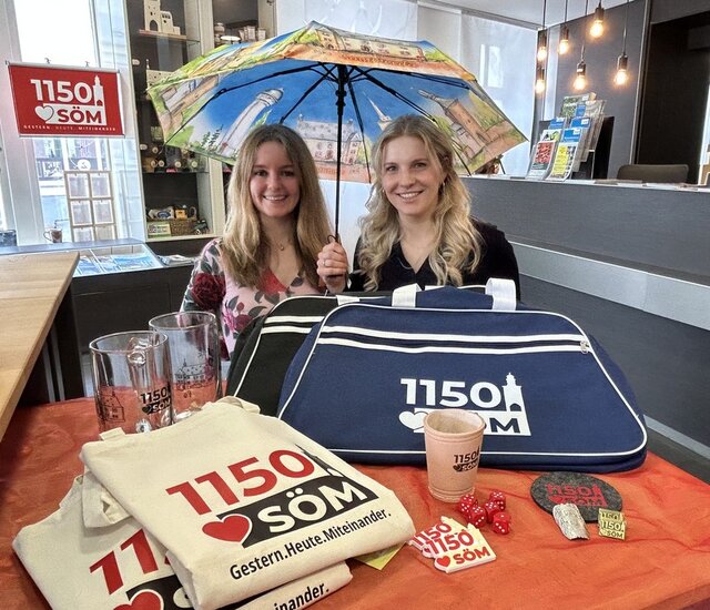 Ein kleiner Tisch in der Tourist-Information ist bedeckt mit Souveniers: Taschen, Beutel, Würfelbecher mit Würfeln, Pins, Stocknägel Untersetzer und Schirmen. Dahiner die Gesicher der zwei Mitarbeiterinnen, die einen Schirm mit Sömmerda-Motiven über sich aufgespannt haben.