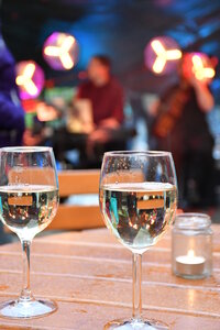 Zwei Gläser Weißwein stehen auf einem Holztisch im Vordergrund, daneben ein kleines Windlicht. Im unscharfen Hintergrund sorgen warme Lichter und sitzende Gäste für eine stimmungsvolle, genussvolle Atmosphäre.
