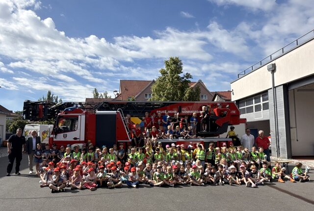 Die Schulstarter sowie Stadtbrandmeister Schönfeld (l.), Bürgermeister Hauboldt (2. v. l.) und Matthias (2. v. r.) und Helmut Schulze (r.).. Das Feuerwehrfahrzeug mit der Drehleiter steht außerhalb der Garage auf dem Gelände der Feuerwache. Davor haben sich die Kinder mit ihren Zuckertüten stehend oder sitzend plaziert. Einige stehen auch auf der Plattform an der Drehleiter. Jedes Kind hält seine Zuckertüte in den Händen.