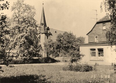 Historische Schwarz-Weiß-Aufnahme aus Frohndorf mit Blick auf eine Kirche mit hohem Kirchturm, umgeben von Bäumen, Wiese und einem Wohnhaus im Vordergrund.