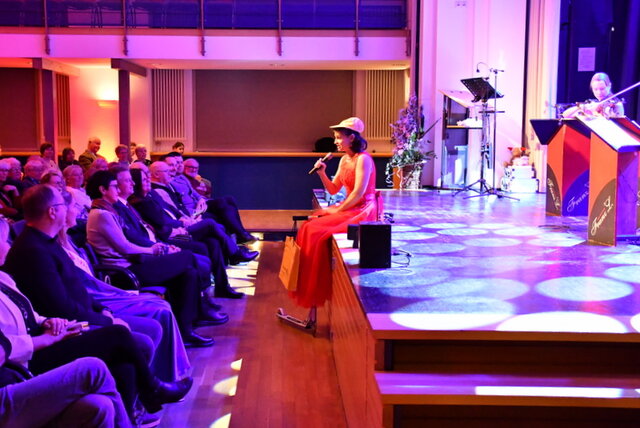 Eine Frau mit rotem Kleid und Basecap sitzt am Rand der Bühne im Volkshaus und singt dem Publikum im Saal zugewandt, das von seinen Stuhlreihen aus auf die Frau und die Bühne blickt. Auf der Bühne hat die Kamera zwei Notenpulte, an denen Franz 'L geschrieben steht. Vom kleinen Orchester wird im Bildausschnitt eine Geigen-Spielerin im roten Kleid gezeigt, die gerade auf ihrem Instrument spielt.
