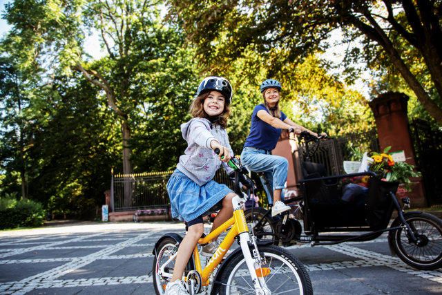 Ein kleines Mädchen sitzt auf seinen gelben Fahrrad, die Mutter dahinter