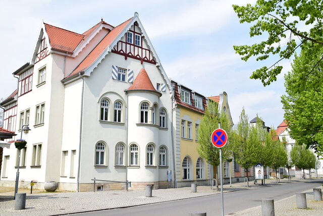 Straßenansicht des Volkshauses Sömmerda in der Weißenseer Straße. Das helle Gebäude mit roten Dachziegeln und verzierten Giebeln liegt an einer gepflasterten Straße, die von Bäumen gesäumt ist. Im Vordergrund steht ein Verkehrsschild mit Halteverbot.