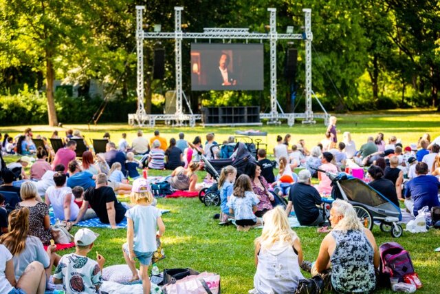 Zahlreiche Menschen sitzen bei sonnigem Wetter auf Picknickdecken und Campingstühlen auf einer Wiese im Stadtpark Sömmerda und schauen gemeinsam einen Film auf einer großen LED-Leinwand. Im Vordergrund sind Kinder, Familien und Kinderwagen zu sehen.
