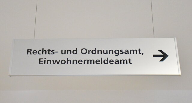 Hinweisschild im Verwaltungsgebäude in der Poststraße 1. Ein rechteckiges Schild aus Edelstahl mhängt von der Decke. Auf dem Schild steht in schwarzer Schrift in der oberen Zeile Rechts- und Ordnungsamt. In der Zeile darunter steht ebenfalls in schwarzer Schrift geschrieben das Wort Einwohnermeldeamt. Ein Pfeil am rechten Ende des Schildes gibt die Richtung vor - nach rechts -, in welcher man zum Einwohnermeldeamt kommt.