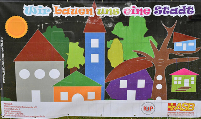 Das Banner für die Kinderstadt, auf dem unter anderem bunte Gebäude, ein Baum, die Logos von ASB und KJP, die Anschrift vom ASB sowie der Schriftzug "Wir bauen uns eine Stadt" im Großformat aufgedruckt sind.
