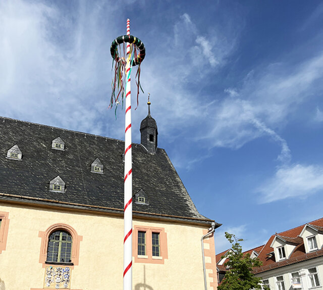 Blick von unten auf den Maibaum, vor dem Rathaus Sömmerda. Der Mast des Maibaums ist in Rot-Weiß, den Farben der Stadt Sömmerda gehalten. Der große Ring an der Spitze des Maibaums ist mit Grün und buten Bändern umwickelt. Ein blauer Himmel mit weißen Wolken schwebt darüber.