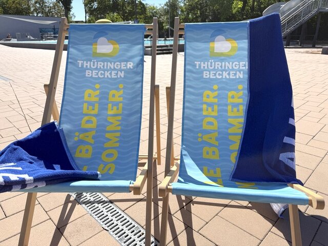 Zwei mit hellblauem Stoff bespannte Liegestühle stehen vor dem Schwimmerbecken im Stadtbad Sömmerda. Auf den Liegestühlen steht in gelber Schrift das Motto der Bäderaktion des Tourismusverbandes Thüringer Becken. Auf einem Liegesrtuhl liegt ein zusammenfaltetes blaues Badetuch. Über dem anderen Liegestuhl hängt ein auseinandergefaltetes Badetuch.
