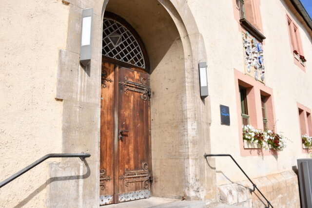 Ein Teil der Vorderseite des Rathauses mit beidseitigem Treppenaufgang und geschlossener Rathaustür. In den Fenstern rechts von der Tür stehen Blumenkästen mit Sommerblühern. Rechts oberhalb der Rathaustür sind verschiedene Wappenreliefs an der Rathaus-Fassade angebracht.