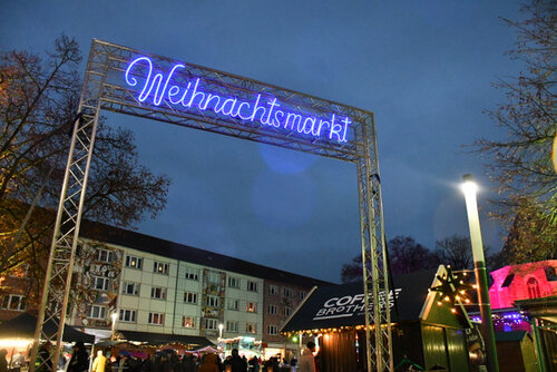 Im Eingangbereich des Weihnachtsmarktes leuchtet der Schriftzug Weihnachtsmarkt in blauer Farbe. Dahinter wird der Blick frei auf verschiedene Hütten und Besucher des Weihnachtsmarktes sowie Häuser, die den Obermarkt begrenzen, freigegeben.