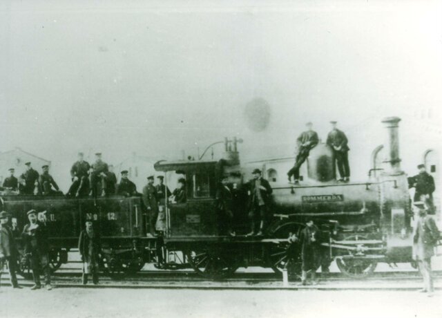 Zahlreiche Männer stehen auf einer alten Eisenbahn aus den 1880er-Jahren und schauen in die Kamera