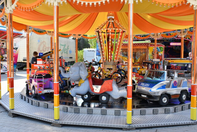Ein farbenfrohes Kinderkarussell mit kleinen Feuerwehr-, Polizei- und Elefantenfahrzeugen dreht sich auf dem Frühlingsfest. Ein Kind sitzt in einem der Wagen. Das Karussell ist mit orange-gelbem Stoff überdacht und festlich geschmückt. Im Hintergrund sind weitere Fahrgeschäfte und bunte Buden zu sehen.