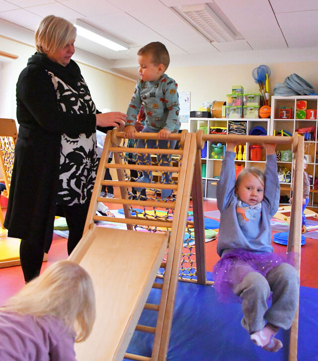 Ein kleines Klettergerüst mit Rutsche aus Holz steht im Sportraum der Kita. Eine Erzieherin hält ein Kind am Kletternetz. Ein weiteres Kind schaukelt an dem Gerüst.