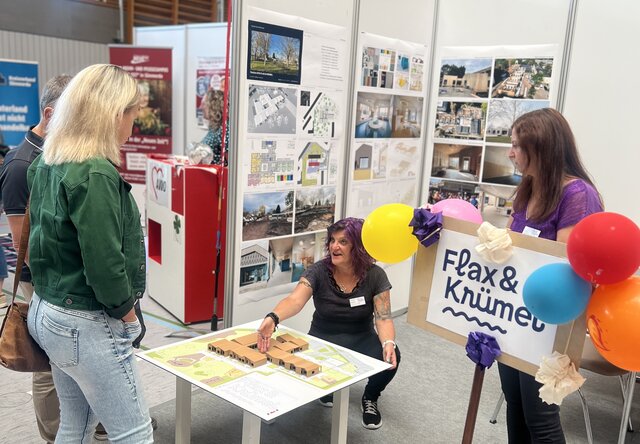 Städtischer Messestand der Kita „Flax & Krümel“ auf der SÖM 2026 Eine Besucherin spricht am Messestand der städtischen Kita „Flax & Krümel“ mit zwei Mitarbeiterinnen. Auf einem Tisch ist ein 3D-Modell des Kita-Neubaus zu sehen, im Hintergrund informieren Schautafeln über das Bauvorhaben.
