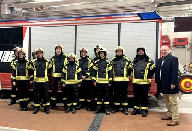 Einsatzkräfte der Feuerwehr in ihrer neuen Kleidung mit Bürgermeister Ralf Hauboldt. Elf Feuerwehrfrauen und -männer stehen in zwei Reihen im der Gerätehalle der Feuerwache vor einem Feuerwehr-Einsatzfahrzeug. Sie alle tragen die die neue dunkelblaue Einsatzkleidung mit neongrünen Reflektoren und ihren Schutzhelm. Der Bürgermeister steht rechts neben der Gruppe.