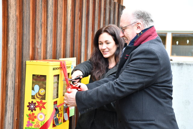 Anne Schmidt, Leiterin der Bibliothek und Bürgermeister Ralf Hauboldt zerschneiden eine rote Schleife und eröffnen offziell den gelben Bienenfutterautomaten im Rosengarten der Stadt- und Kreisbibliothek