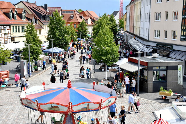 Die Marktstraße in Sömmerda an einem belebten Veranstaltungstag: Zahlreiche Menschen schlendern bei sonnigem Wetter entlang der mit Bäumen gesäumten Straße. Links und rechts sind Stände, Pavillons und Marktbuden aufgebaut. Im Vordergrund ist ein rot-blaues Kinderkarussell zu sehen. Die umliegenden Gebäude sind geschmückt mit Schildern, Markisen und Schaufenstern. Die Atmosphäre ist lebendig und einladend.