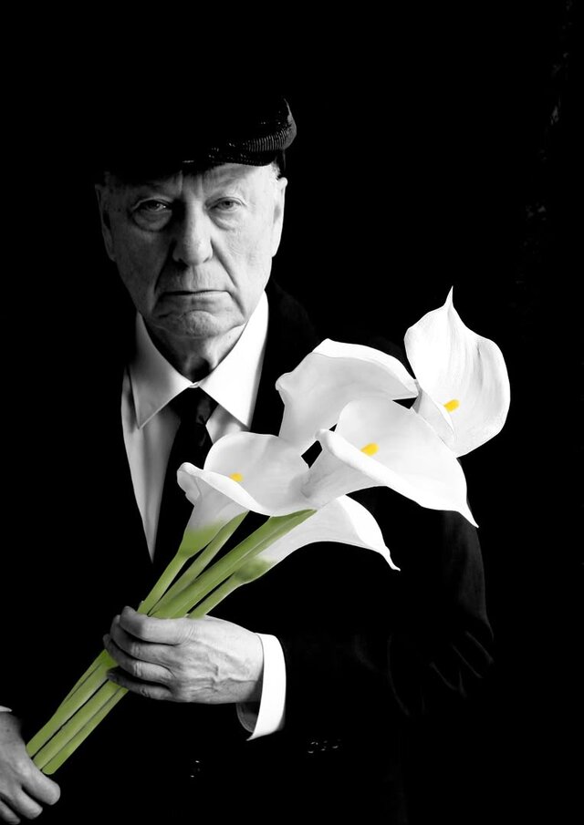 Schwarz-weiß Aufnahme von Gerd Dudenhöffer in einem schwarzen Anzug vor schwarzem Hintergrund. In der Hand hält er fünf Callas.