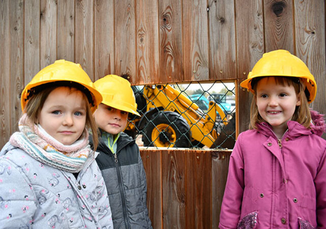 Drei Kita-Kinder mit gelben Bauhelmen stehen vor einem Bauzaun aus Holz. Der Zaun hat ein Sichtfenster in Kopfhöhe der Kinder. Durch das Fenster sieht man ein Baufahrzeug.