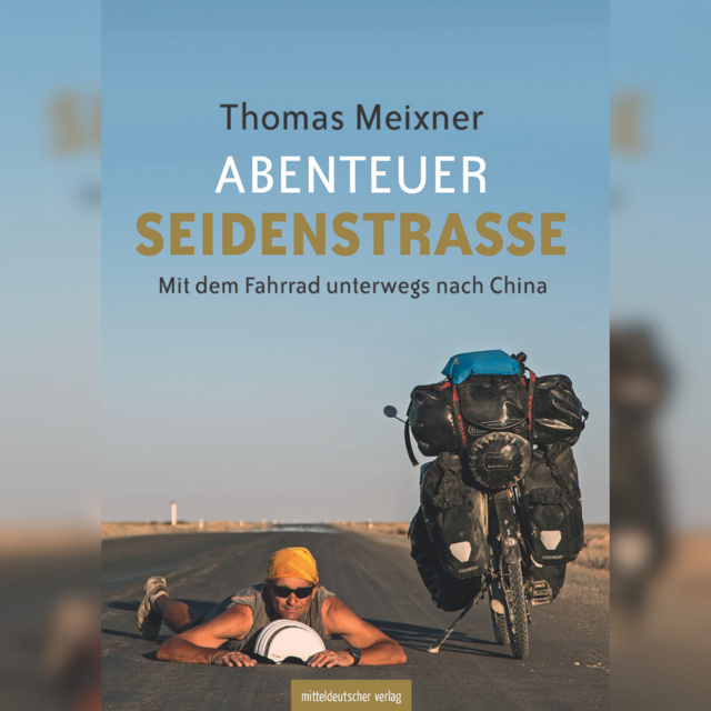Auf dem Bild ist der Buchtitel "Abenteuer Seidenstraße" von Thomas Meixner zu sehen, er liegt auf der Straße, neben ihm steht sein Fahrrad mit dem Gepäck