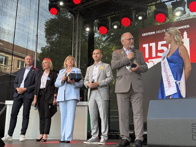 Auf der Bühne beim Stadtjubiläum stehen der Landrat sowie die stellvertretende Bürgermeisterin von Kedainiai, die Dolmetscherin und der Oberbürgermeister von Böblingen in einer Reihe. Leicht nach vorn gerückt steht Sömmerdas Bürgermeister mit der Sömmerdaer Waidprinzessin. Sie trägt ein langes blaues Kleid, eine weiße Schärpe mit roter Schrift sowie ein Diadem im blonden Haar. Bürgermeister und Waidprinzessin führen ein kurzes Gespräch über Mikrofon.