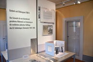 Ein kleiner Raum mit der Ausstellung der vier Entwürfe für das Mahnmal. Zu sehen ist der Siegerentwurf. Dahinter an einer Tafel steht der Titel der Ausstellung: Gedenk- und Bildungsort Tor 8: Vier Entwürfe für ein künstlerisch gestaltetes Mahnmal zur Erinnerung an die weiblichen jüdischen Häftlinge des KZ Außenlagers Sömmerda 1944-1945.