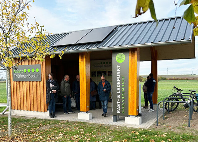 Ein überdachter Holzbau mit Solarpaneelen auf dem Dach. An dem Holzbau sind links und rechts Tafeln angebracht. Links mit dem Logo des Zweckverbandes Allianz "Thüringer Becken". Rechts mit dem Hinweis auf den rast- ud Ladepunkt Großbrembach. Zwei Fahrräder stehen am Holzbau. Unter dem Dach stehen mehrere Leute.