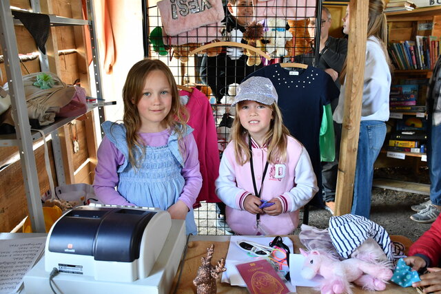 Zwei Mädchen stehen hinter der Verkaufstheke im Spittelladen der Kinderstadt Sömmerda. Auf dem Tisch liegen Spielwaren, Kinderaccessoires und eine Spielkasse. Im Hintergrund sind Kleidung, Bücher und weitere Kinder sowie Erwachsene zu sehen. Kinder können hier ihre erspielten „Unstruttaler“ einlösen.