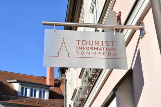 Ein Ausleger am Gebäude der Tourist-Information. Dabei handelt es sich um ein Schild, das an einer Stande aus Edelstahl angebracht ist. Auf dem flachen Schild aus Edelstahl steht in roter Schrift Tourist-Information Sömmerda. Ebenfalls in rot gehalten befindet sich eine Silhouette von Sömmerda auf dem Schild.