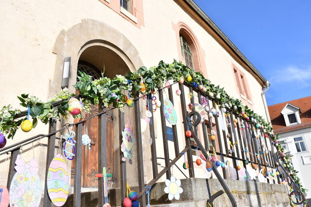Das Geländer am beidseitgen Treppenaufgang zum Rathaus-Eingang ist am Handlauf mit einer grünen Girlande umwunden. Darin hängen Ostereier in verschiedener Ausführung: bemalt oder aus starkem Papier und bunt bemalt. Die Sonne scheint und der Himmel leuchtet blau.