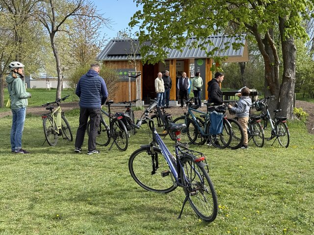 Mehrere Radfahrerinnen und Radfahrer stehen mit ihren Fahrrädern am neu eröffneten Radler-Rastplatz in Leubingen. Im Hintergrund ist die überdachte Holzstation mit Solarmodulen zu sehen.