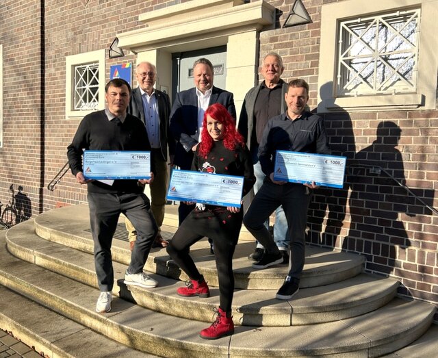 Nach der Scheckübergabe vor dem SEV-Gebäude. Die schs Beteiliten der Scheckübergabe stehen gestaffelt auf der Treppe zum SEV-Gebäude. Die Sonne scheint. Die Vertreter der drei Vereine präsentieren die symbolischen Schecks. Alle sechs schauen lächeln in die Kamera.
