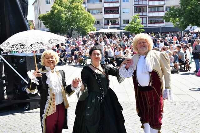 Drei in historischen Gewändern verkleidete Menschen schauen in die Kamera. Hinter ihnen sieht man viele Leute an Tischen sitzen und es herrscht ein buntes Treiben.