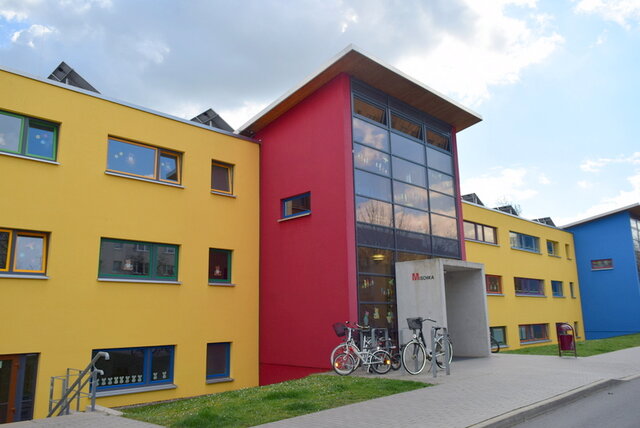 Das Gebäude, das die städtische Kita "Mischka" sich mit der Evangelischen Grundschule teilt. Ein neuerer Bau mit vorgezogenem und an der Frontseite komplett verglastem Eingangsportal. Das dreistöckige Gebäude ist farblich in Gelb und Rot gehalten. Davor sind eine Zufahrt und eine schmale Rasenfläche zu sehen.