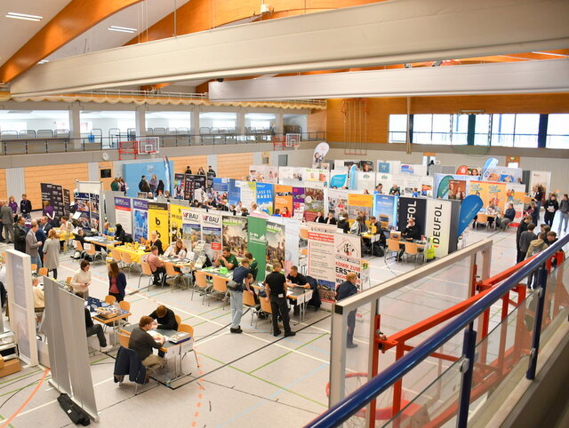 In der großen und hellen Unstruthalle - eine Sport- und Mehrzweckhalle - stehen in mehreren Reihen Stände verschiedener Unternehmen und Institutionen nebeneinander. Hinter den Tischen mit den Werbebannern der Unternehmen sitzen Firmenvertreterinnen und Firmenvertreter. Vor den Tischen - allerdings nicht vor allen, sitzen junge Leute. Sie unterhalten sich mit den Firmenvertreterinnen und Firmenvertretern. 