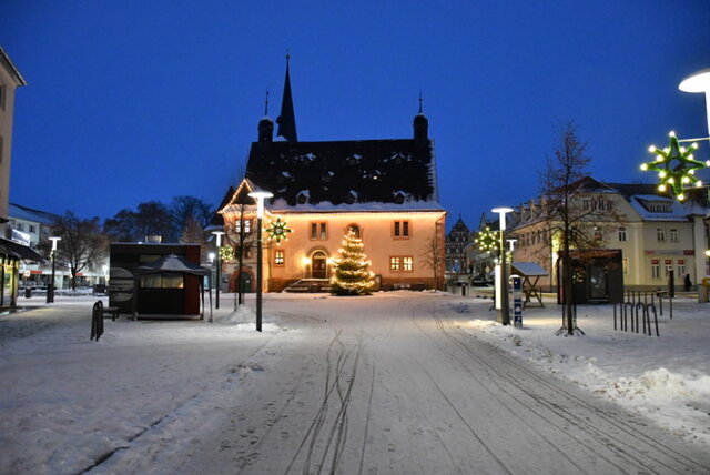 Das mit Lichtkern erleuchtete Rathaus in der Dämmerstunde mit dem schöm beleuchtetwen Weihnachtsbaum davor. Wir blicken aus der Marktstraße aus einigem Abstand direkt auf Rathaus und Weihnachtsbaum. An den Straßenlampen hängen leuchtende Sterne. Es liegt eine leichte Schneedecke.