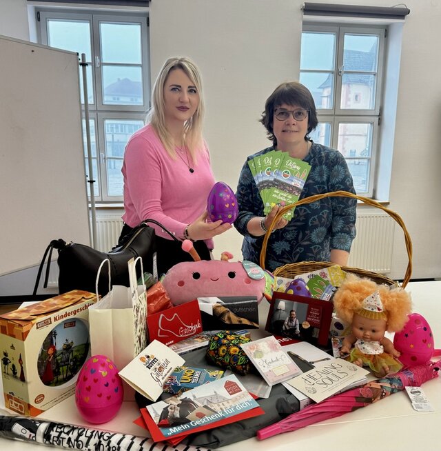 Zwei Frauen stehen hinter einem Tisch. Eine der Frauen hält präsentier ein größeres farbiges Osterei aus Plastik. Es ist eines von den Eiern, die in den Geschäften versteckt wurden. Die andere Frau hält einige der Gewinnkarten, die gezogen wurden, in der Hand. Auf dem Tisch vor ihnen liegen bzw. stehen die Gutscheine und Sachpreise, die die Geschäfte gestifet haben.