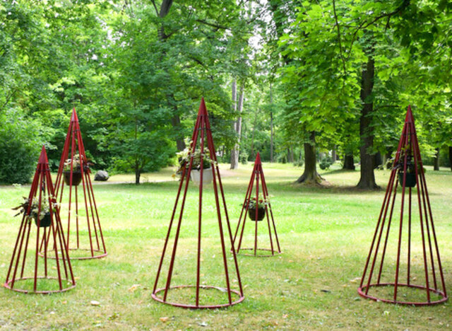Auf einer kurz gemähten Wiese stehend sind für die fünf Blumentipis jeweils mehrere rot lackierte Stangen auf einem ebenfalls roten metallring angeschweißt. Die Stangen führen nach oben spitz zusammen. Die Spitzen werden jeweils von einem kleinen Kegel abgedeckt. Im Inneren des tipiförmigen Gestänges hängt jweils eine mit verschiedenen Blumen bepflanzte Blumenampel. Im Hintergrund stehen mehrere Bäume.