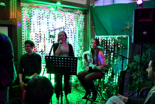 Eine Band aus drei jungen Leuten steht am späten Abend vor einer illuminierten Gaststätte, deren Türen weit geöffnet sind. Die Band spielt Musik und singt, Zuhörer stehen oder sitzen davor.